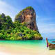 Limestone cliffs covered in green vegetation rising from turquoise water with traditional longtail boat on beach in Thailand